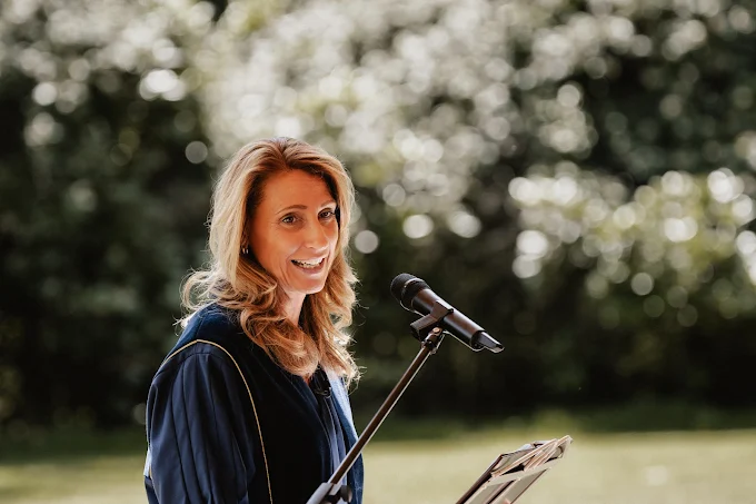 Woman speaking at microphone outdoors with blooming trees