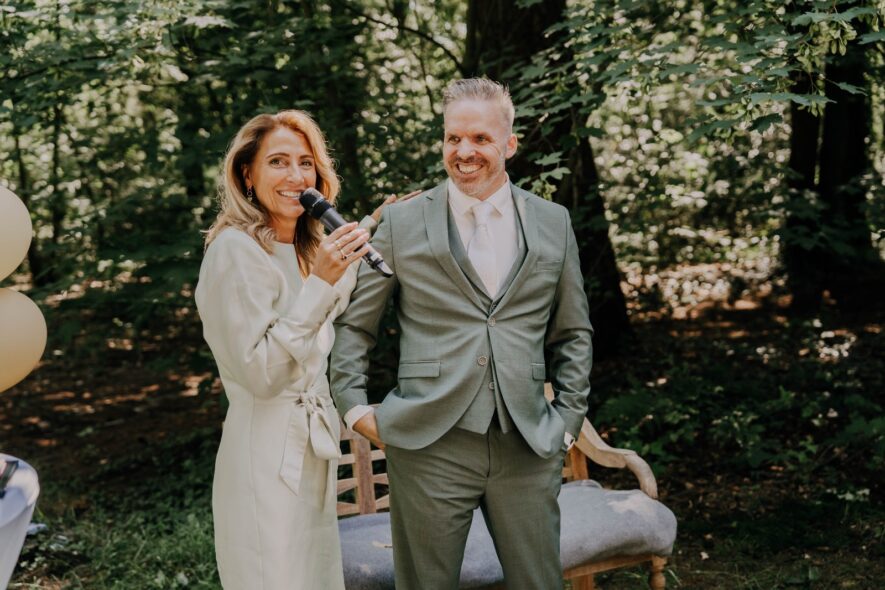 Woman in cream suit holds microphone beside man in sage blazer outdoors